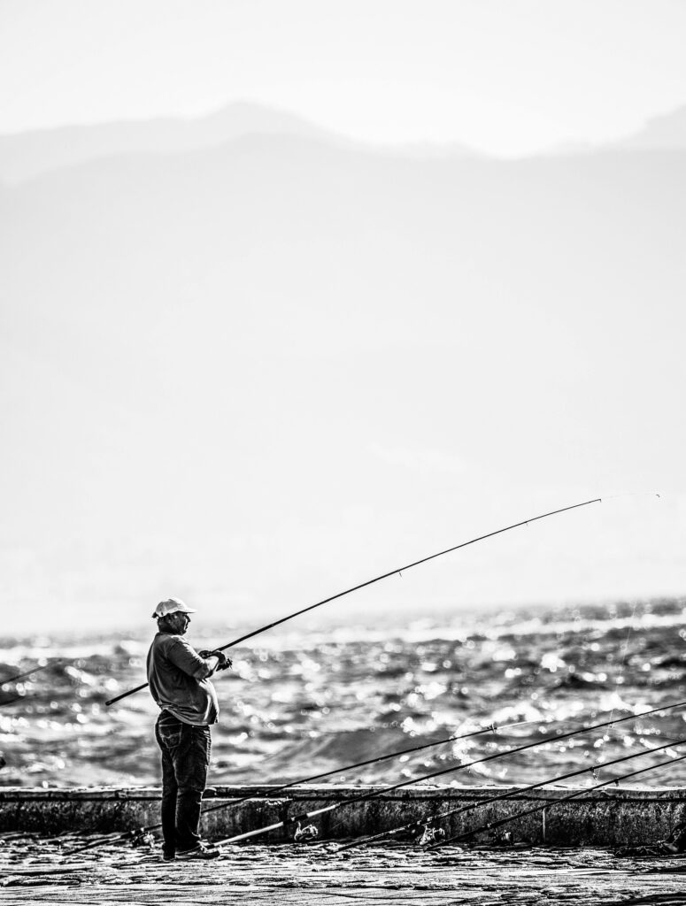 A solitary fisherman with rods stands by the sea under a bright sky.